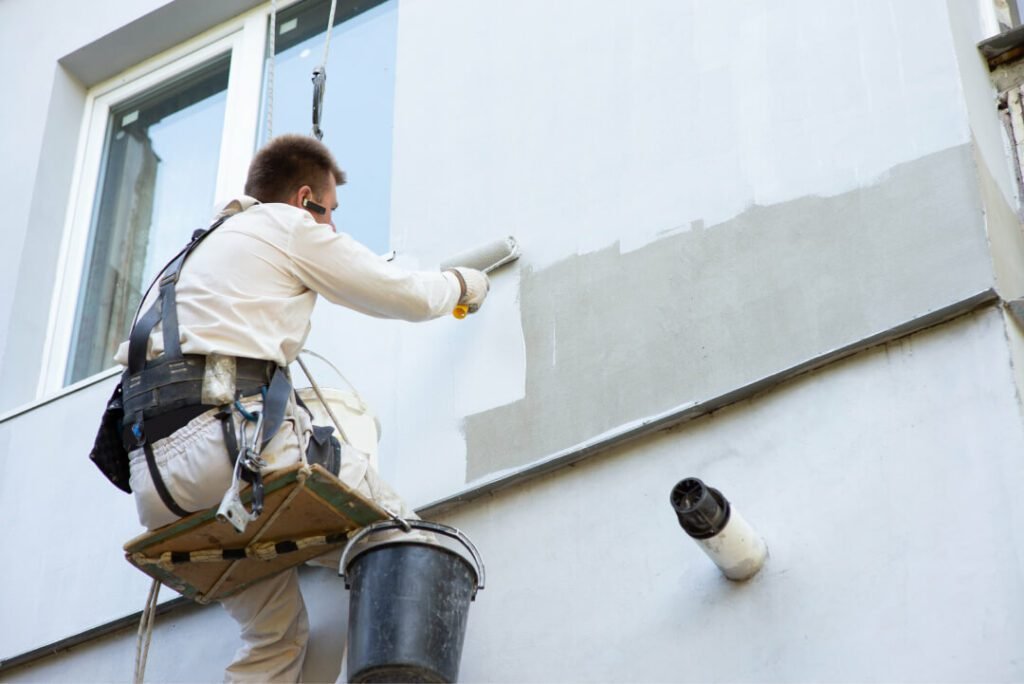 A builder-alpinist painting a building facade with light-colored paint.